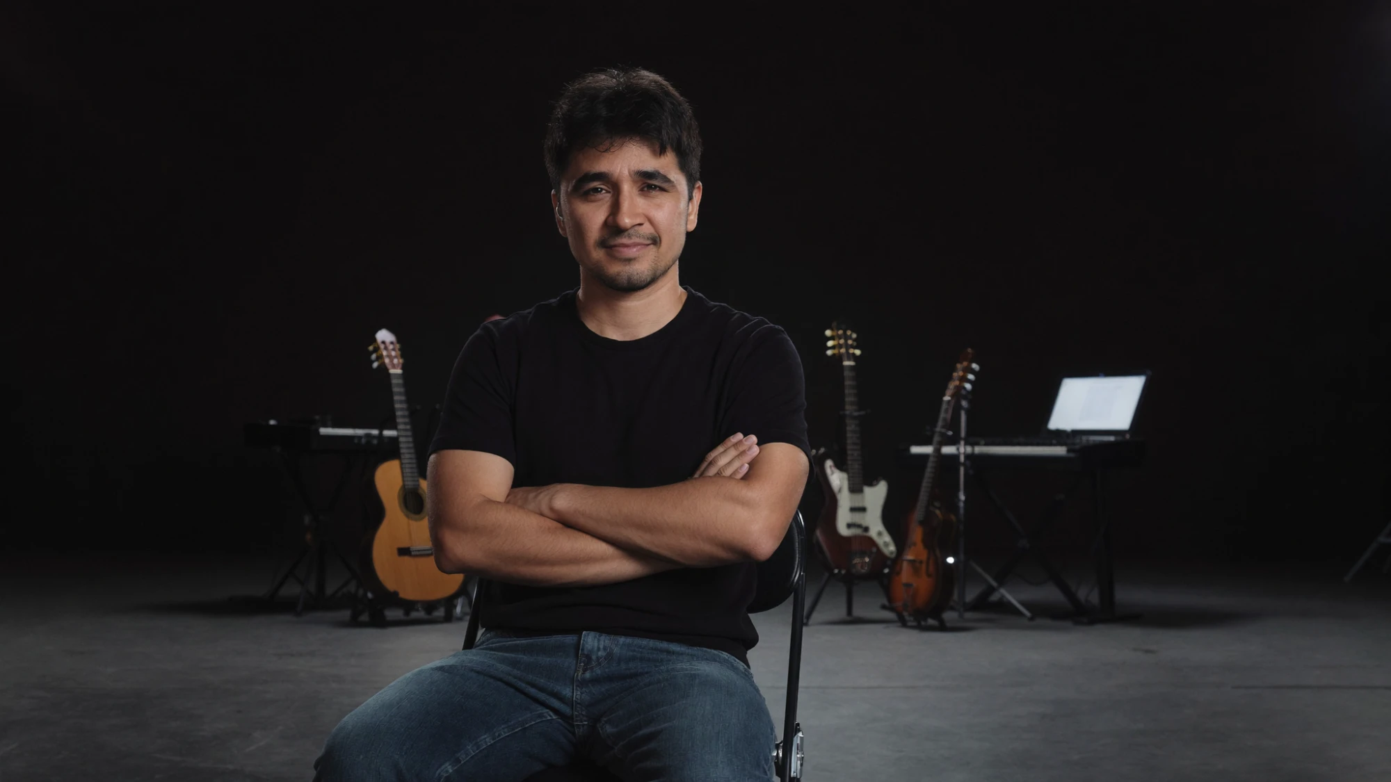 Carlos Jr, sentado de braços cruzados, com violão, guitarra, teclado e um notebook ao fundo